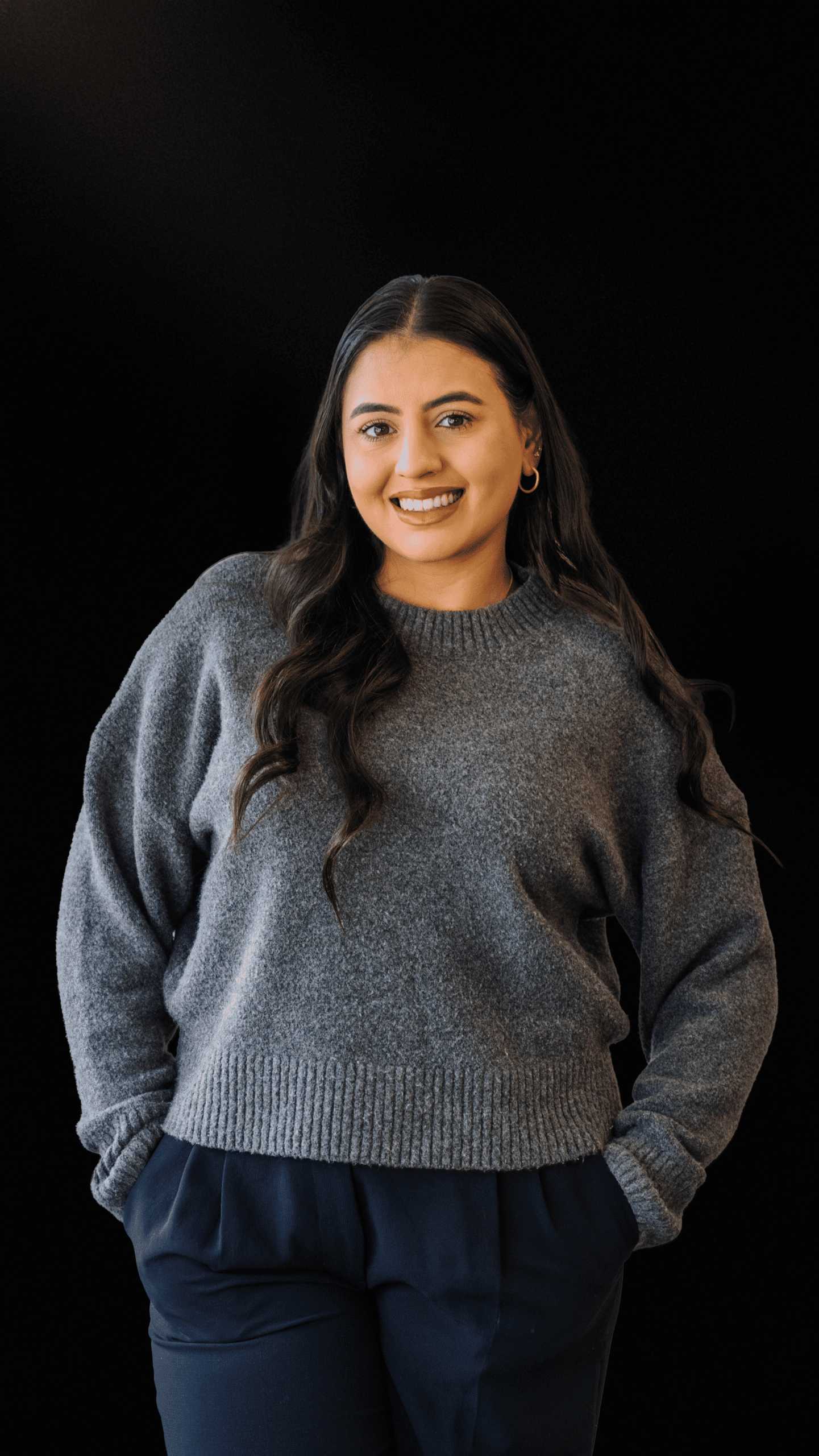 Woman smiling in a grey sweater, standing confidently against a black background.