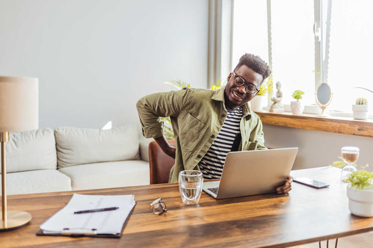 Man sitting at a desk holding her lower back in discomfort with a glass of water next to him